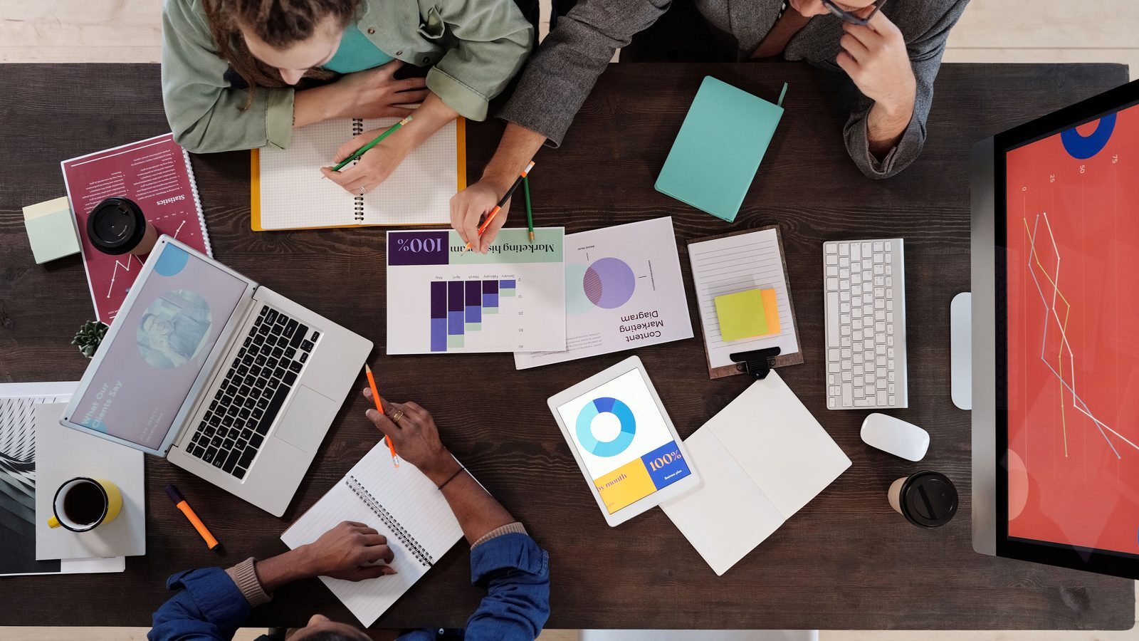 Team arbeitet von oben gesehen an einem Tisch mit Laptop, Tablet, Monitor und ausgedruckten Diagrammen
